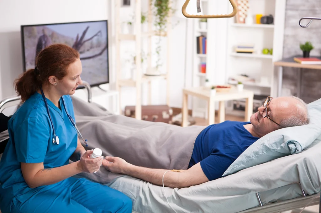 ممرضة تقدم علاجًا بالحقن الوريدي لرجل مسن مستلقٍ في سرير طبي داخل منزله