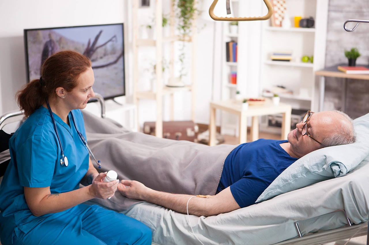ممرضة تقدم علاجًا بالحقن الوريدي لرجل مسن مستلقٍ في سرير طبي داخل منزله